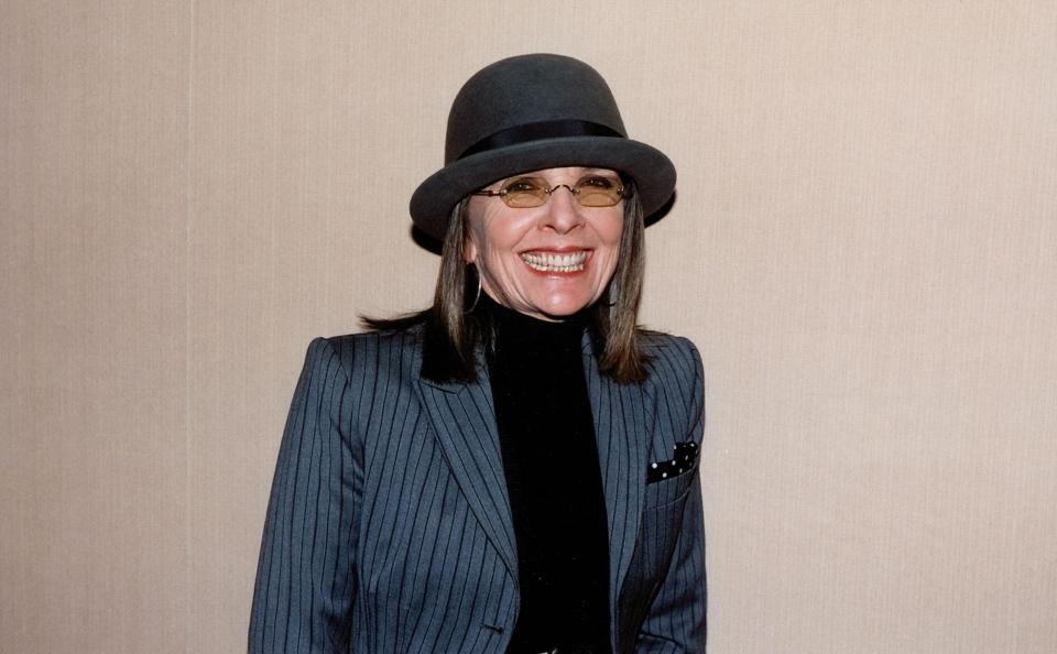 Diane Keaton wearing a pinstripe blazer, black turtleneck, a dark gray fedora, and amber-tinted glasses.