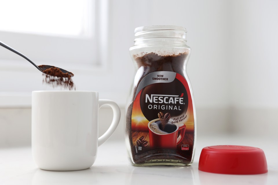 A spoon with Nescafe instant coffee pouring into a white mug next to a jar of Nescafe Original instant coffee.