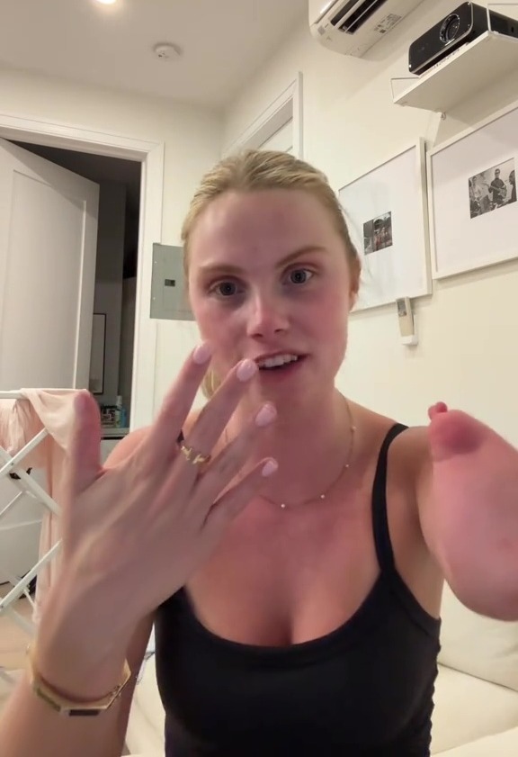 A woman wearing a black tank top and a gold ring and bracelet, shows off her light pink manicured fingernails.