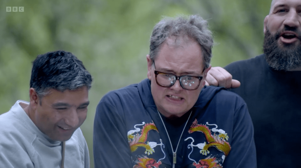 Three men watching something with mixed expressions, one man in glasses wearing a dragon-embroidered hoodie looks distressed.
