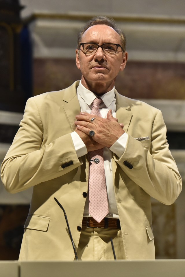 Kevin Spacey addressing the audience, wearing a light tan suit, a white shirt, and a pink patterned tie with a tie clip.
