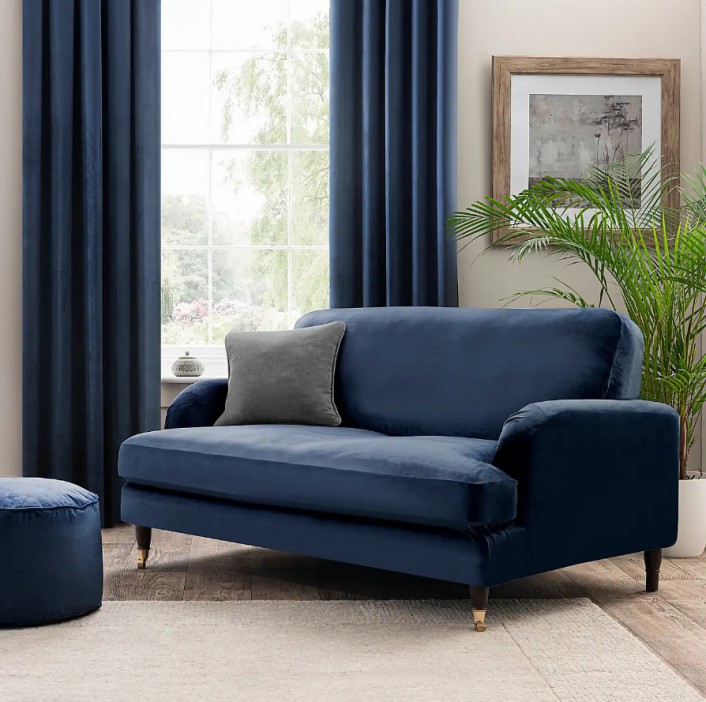 Blue velvet loveseat with a gray throw pillow, a matching ottoman, and blue curtains in a living room.