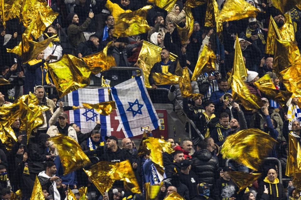 Maccabi Tel Aviv F.C. supporters wave yellow and Israeli flags during a UEFA Europa League match.