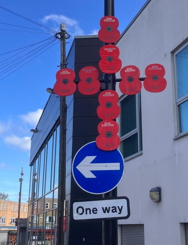 Numerous red poppy displays attached to a lamp post, accompanied by a blue arrow sign indicating "one way."