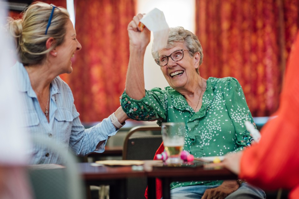 A woman raising a bingo card in the air while laughing, with another woman laughing next to her.