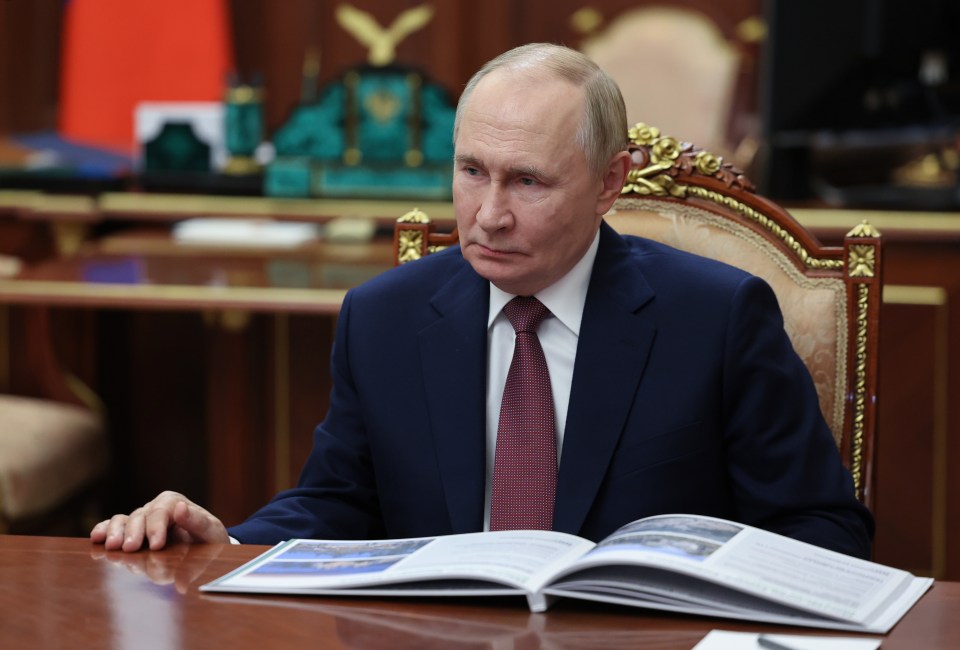 Russian President Vladimir Putin in a suit and tie, seated at a wooden desk with an open book in front of him.