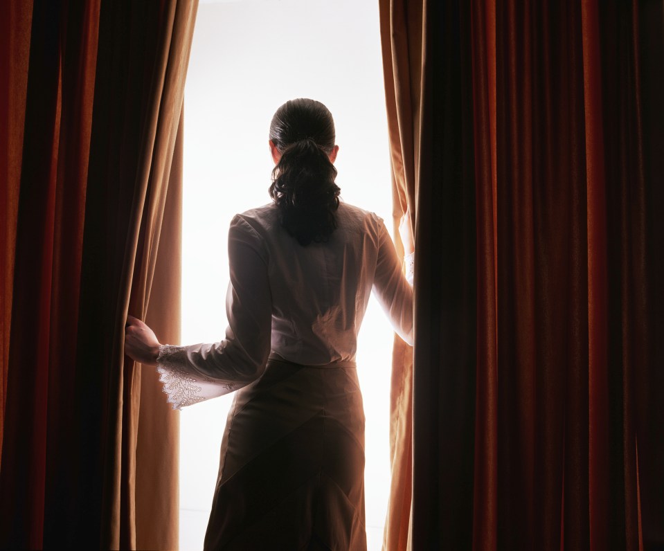 Woman with a ponytail pulling back red curtains to reveal a bright light.