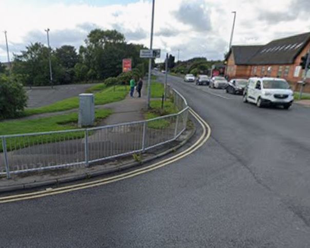 A street view showing a road with traffic, a sidewalk with pedestrians, and an empty parking lot to the left.