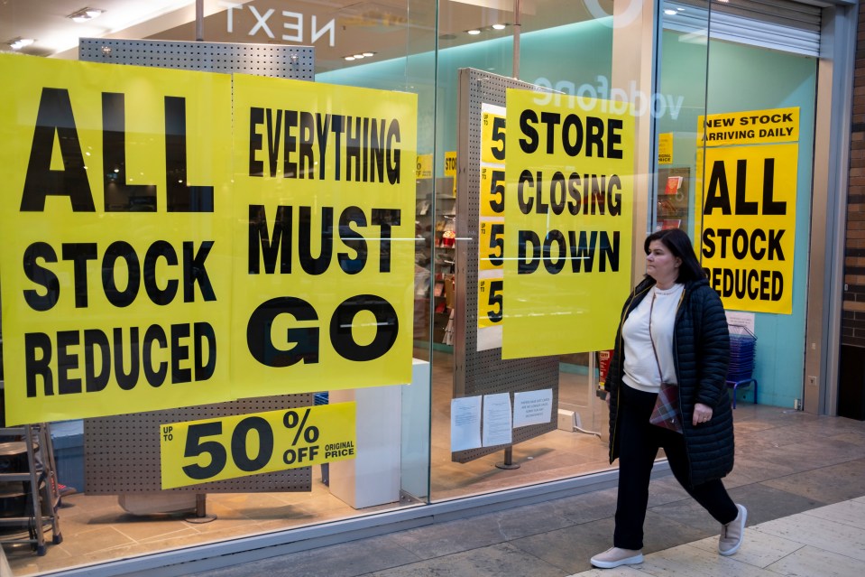 A woman walking past a Paperchase store with "Store Closing Down" and "All Stock Reduced" signs on its windows.