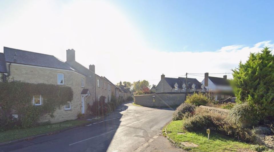 A sunlit street scene in a village, with stone houses lining the road and a large tree on the right.