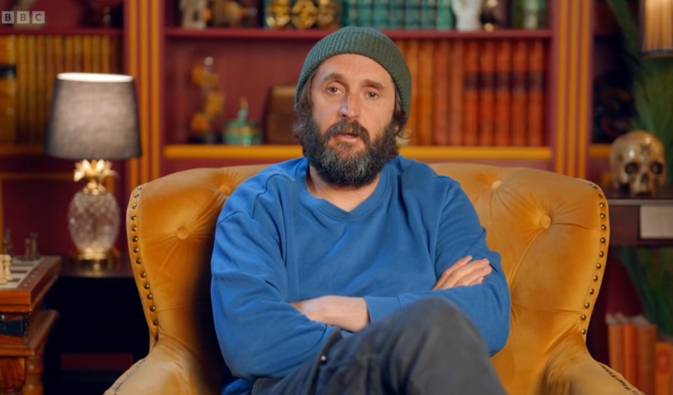 A bearded man in a blue sweatshirt and a green beanie sitting in a yellow armchair.