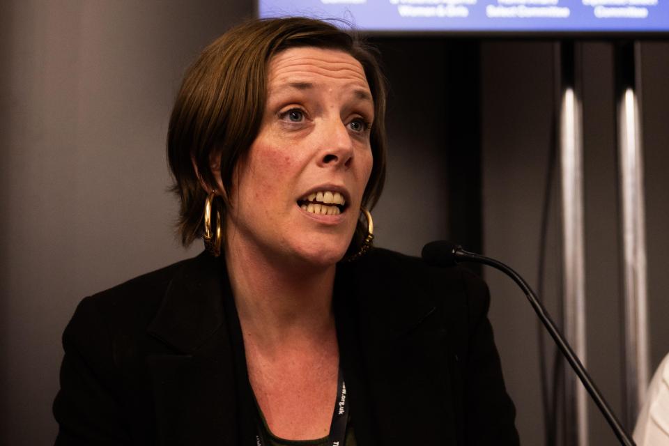 A woman with short brown hair, wearing a black blazer and large hoop earrings, speaking into a microphone.