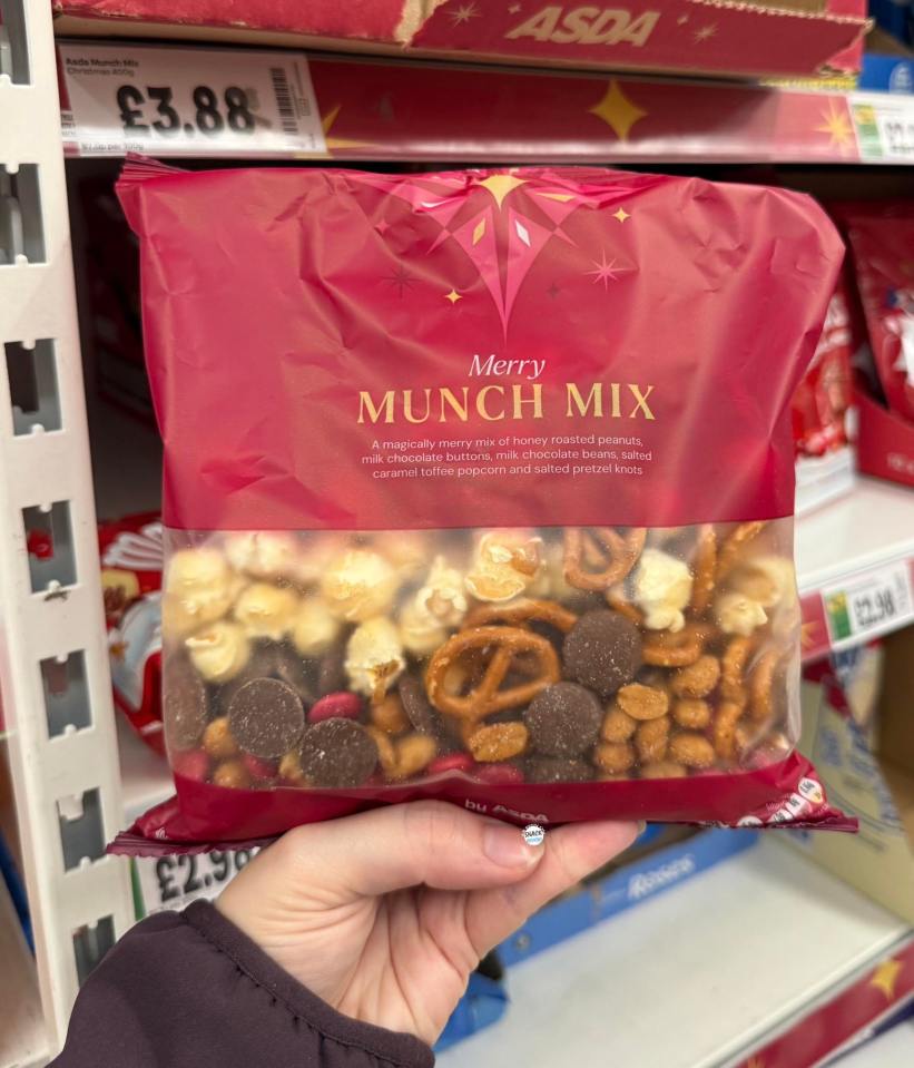 A hand holding a red and clear bag of Asda Merry Munch Mix, displaying popcorn, pretzels, peanuts, and chocolate.