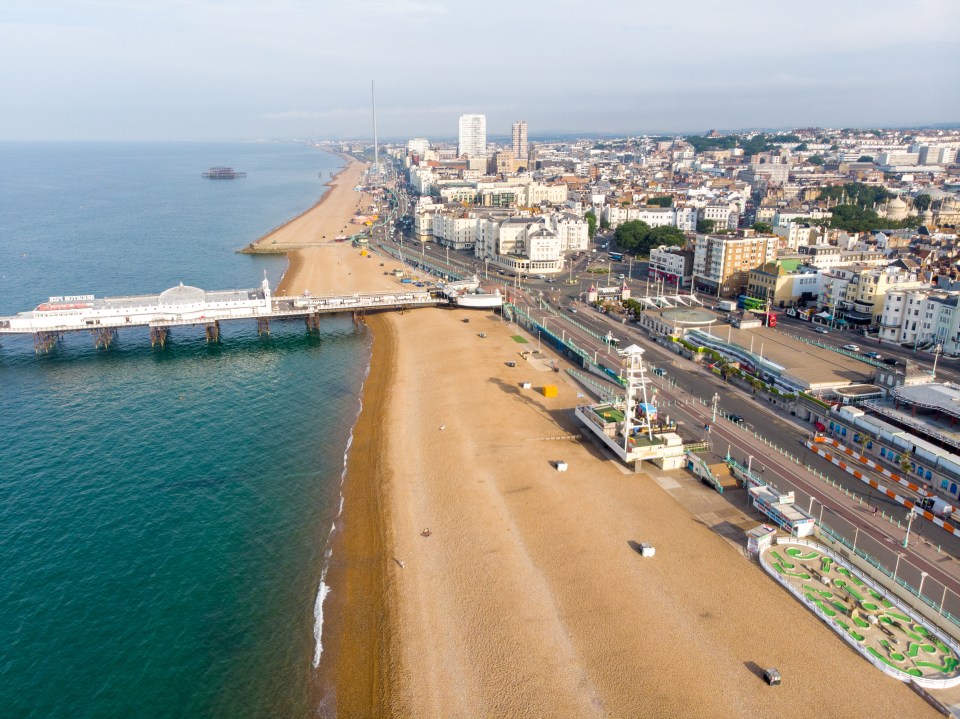 Aerial view of Brighton's beachfront with the Palace Pier extending into the sea, a city skyline in the background, and a wide pebble beach.