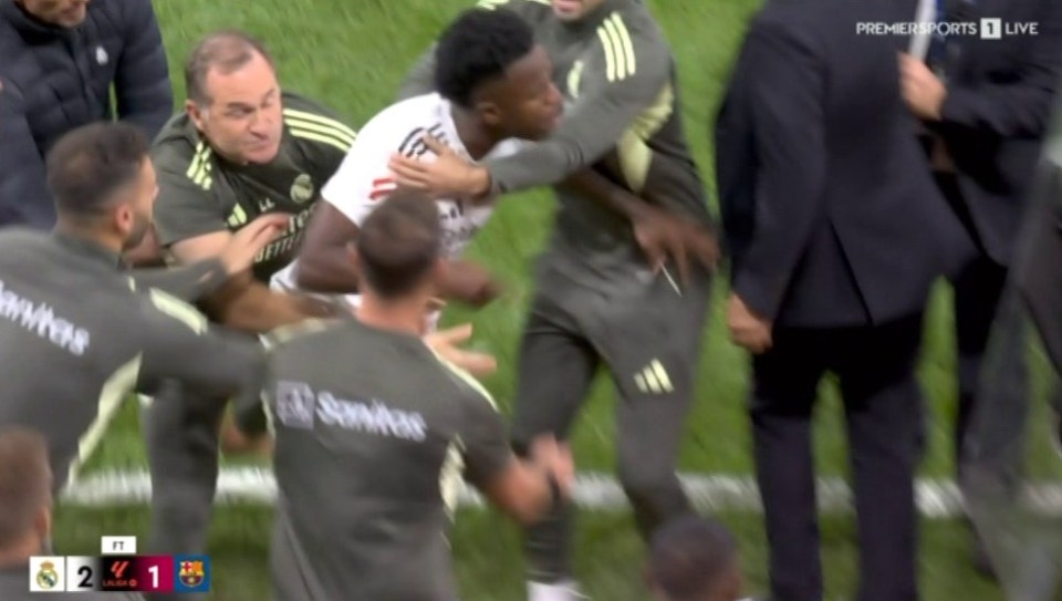 A group of soccer players and staff on a field, some in grey training gear and one in a white and red jersey, in a huddle after a game with a score overlay of Real Madrid 2 - 1 Barcelona.