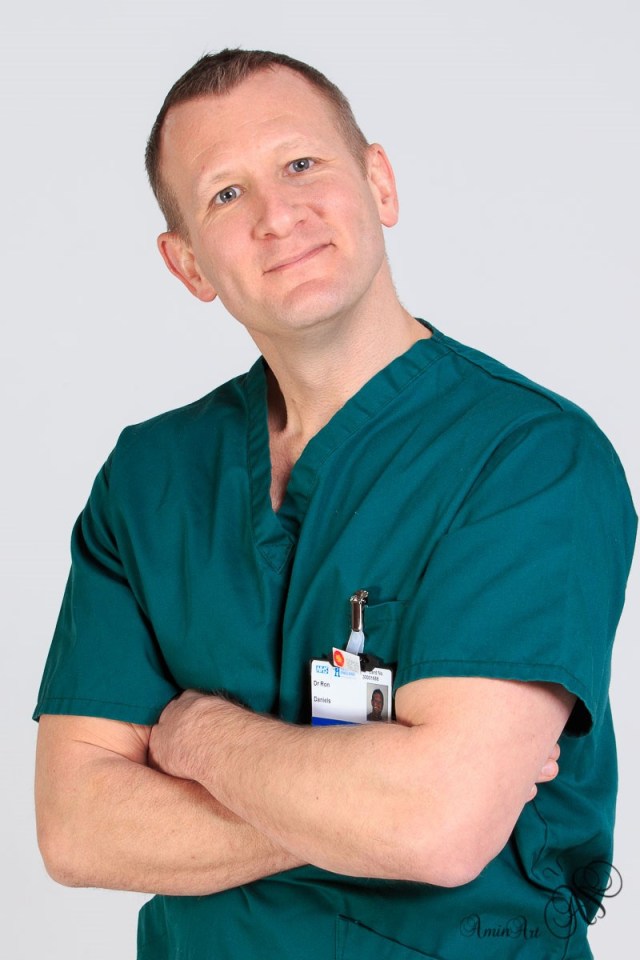 Dr. Ron Daniels, an Intensive Care Consultant, wearing green scrubs and an ID badge, smiling at the camera.