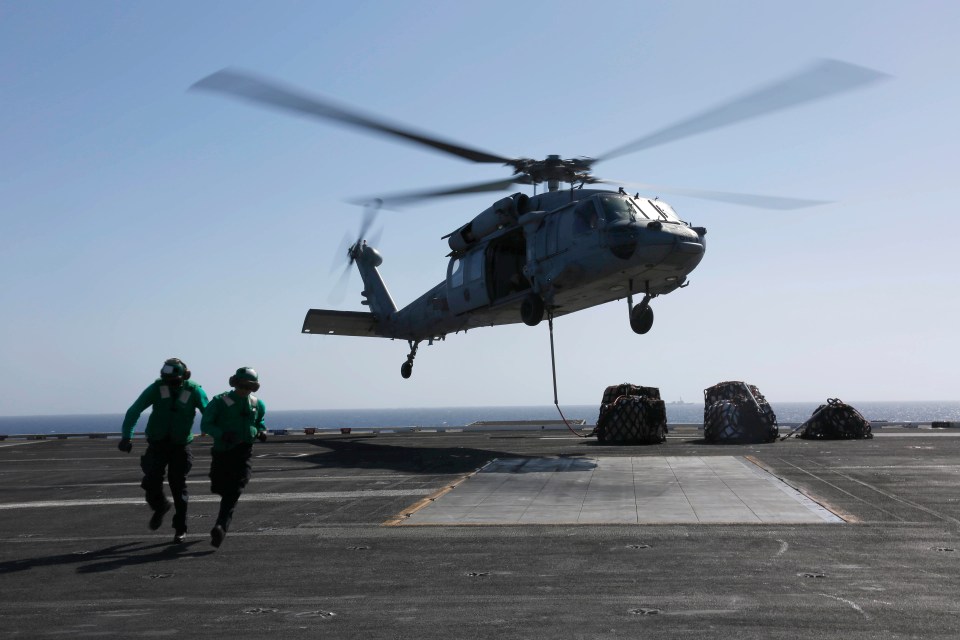 Two naval personnel on a flight deck attach cargo to an MH-60S Sea Hawk helicopter.
