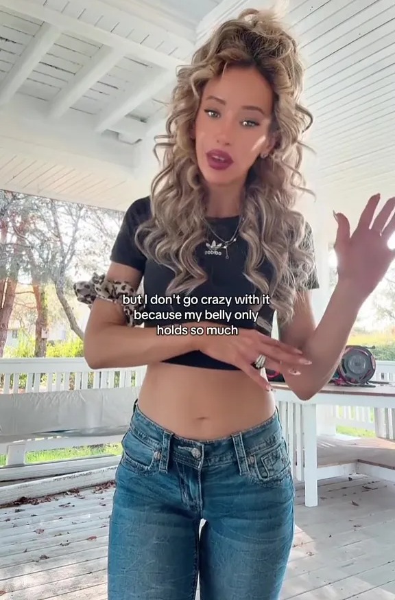 A woman in a black crop top and blue jeans gestures with text overlay, "but I don't go crazy with it because my belly only holds so much."