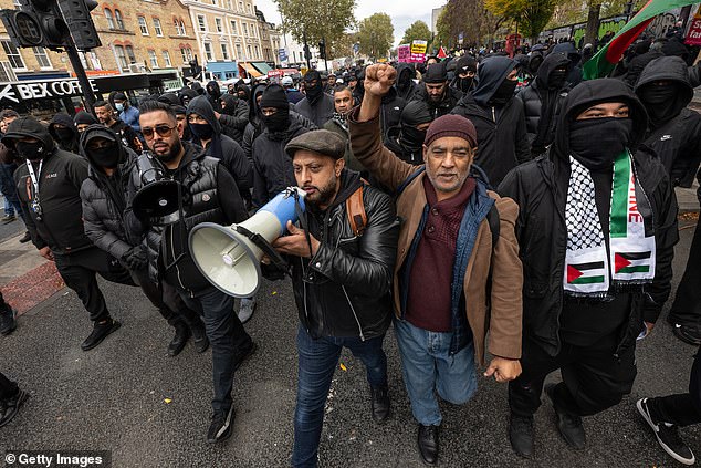 A man raises his fist in the air as the other speaks on a microphone as members of the community take to the streets of east London