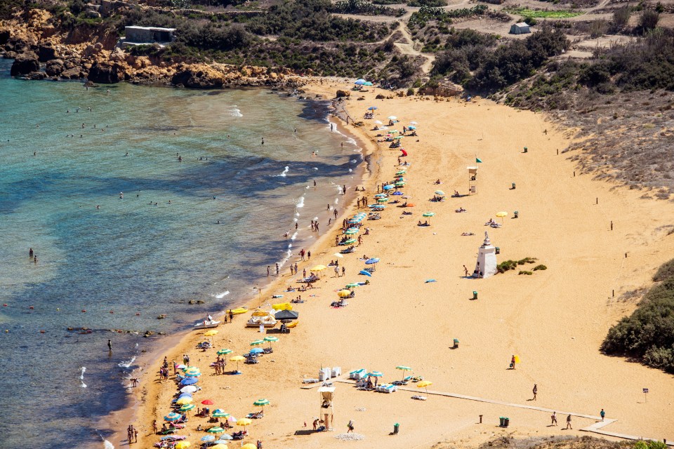 Aerial view of Ramla Bay beach in Gozo, Malta, showing a sandy beach with many people, colorful umbrellas, and clear blue water.