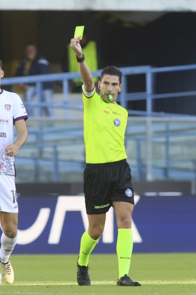 A referee in a neon yellow uniform holds up a yellow card during the Hellas Verona FC vs. Cagliari Calcio Serie A match.