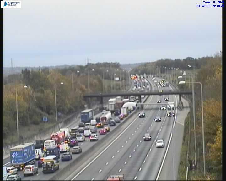 Traffic camera image showing multiple lanes of traffic, with congestion and emergency vehicles in the left lanes, and flowing traffic in the right lanes.