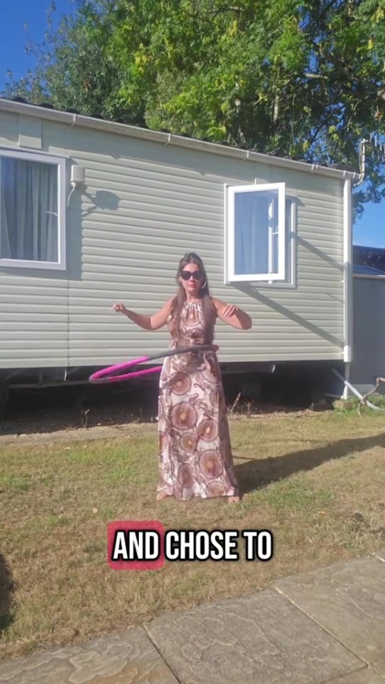 A woman hula-hoops in a yard in front of a caravan.