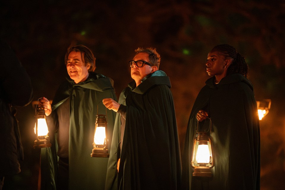 Three people in dark cloaks, each holding a lit lantern, stand in a dark environment.