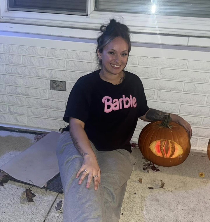A young woman wearing a Barbie t-shirt sits next to a carved pumpkin with glowing eyes.