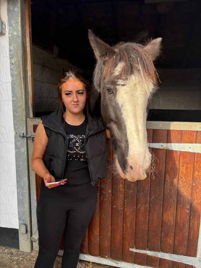 A woman with a tan face stands next to a brown and white horse in a stable doorway.
