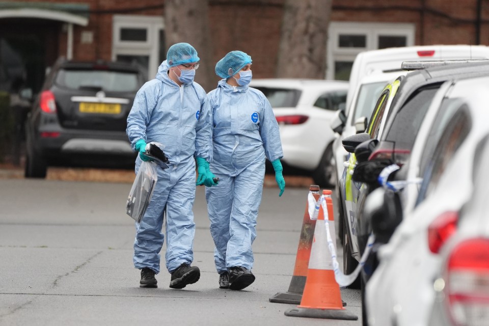 Two forensic officers in light blue protective suits and masks walk on a street with parked cars.