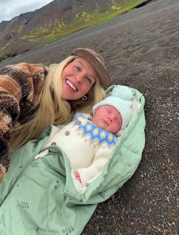 A woman in a brown hat and fluffy jacket smiling at the camera, holding a sleeping infant wrapped in a green blanket, with a mountain in the background.