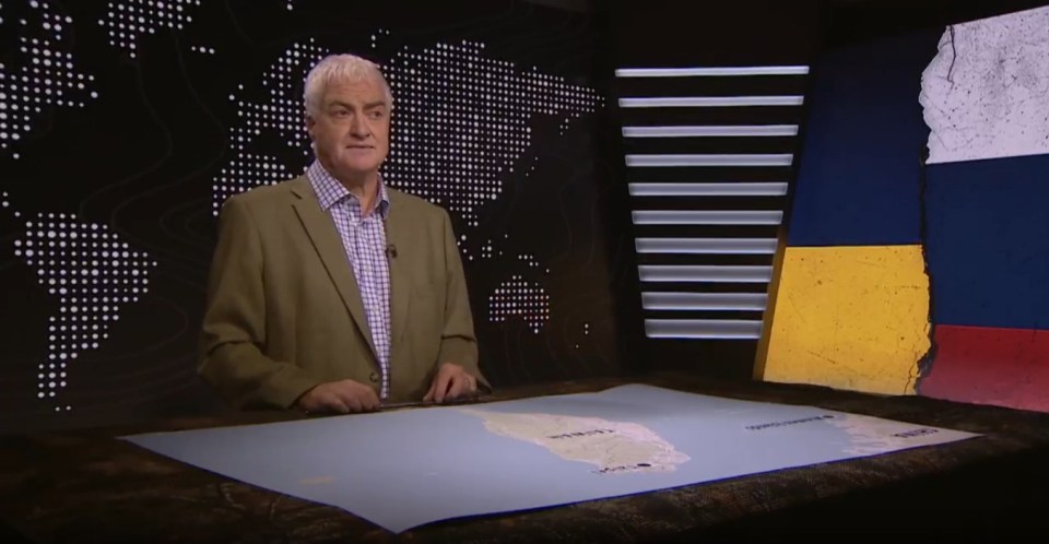 A man in a suit jacket stands in front of a news desk with a map of South America, and a backdrop displaying a world map and a cracked image of the Ukrainian and Russian flags.