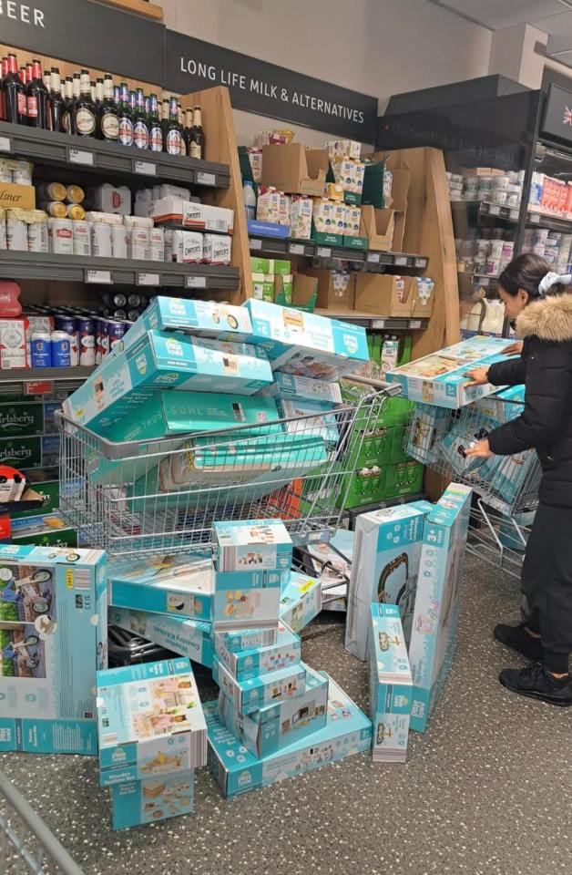 A customer with a shopping cart overflowing with toy boxes, surrounded by more toy boxes on the floor in an Aldi aisle.
