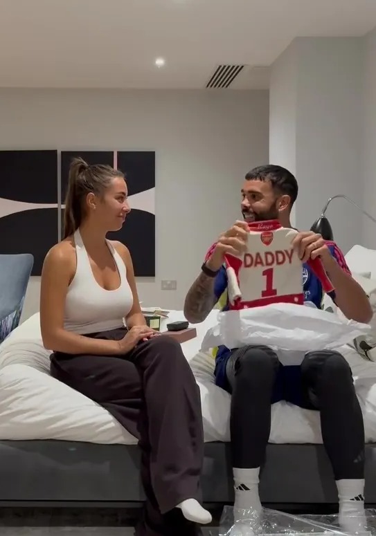 David Raya holding a baby onesie that reads "Daddy 1" while sitting next to a woman.