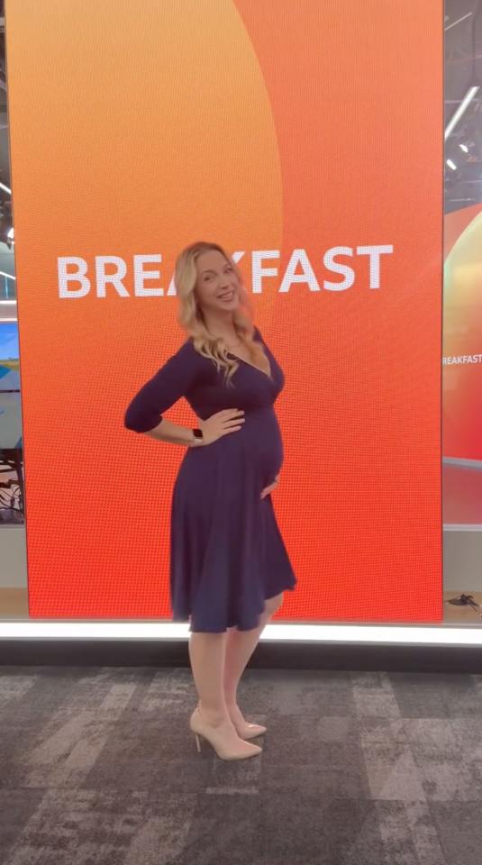 Pregnant woman in a dark blue dress posing in front of a red screen with "BREAKFAST" written on it.