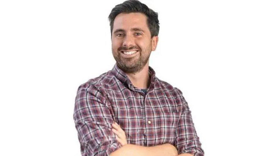 A smiling man with dark hair and a beard, wearing a plaid shirt, with his arms crossed.