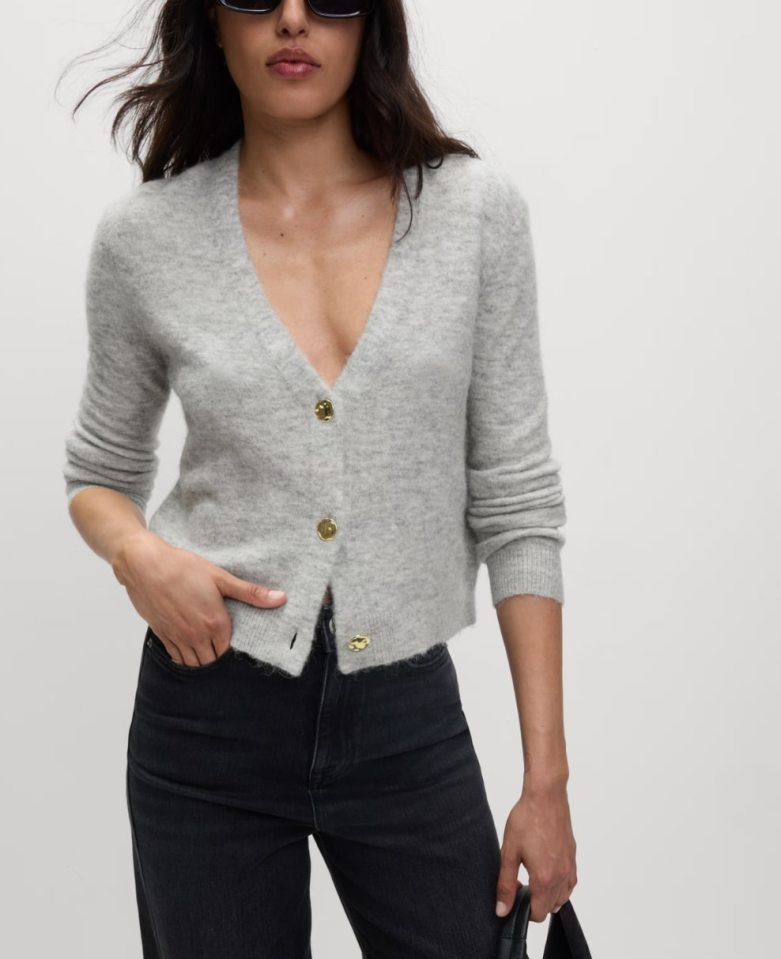 A woman modeling a light grey cardigan with gold buttons and dark wash jeans.