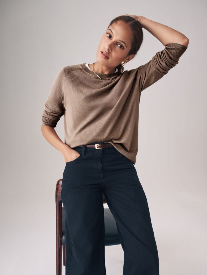 A woman wearing a brown long-sleeved top and dark blue trousers stands with one hand in her pocket and the other on her head.