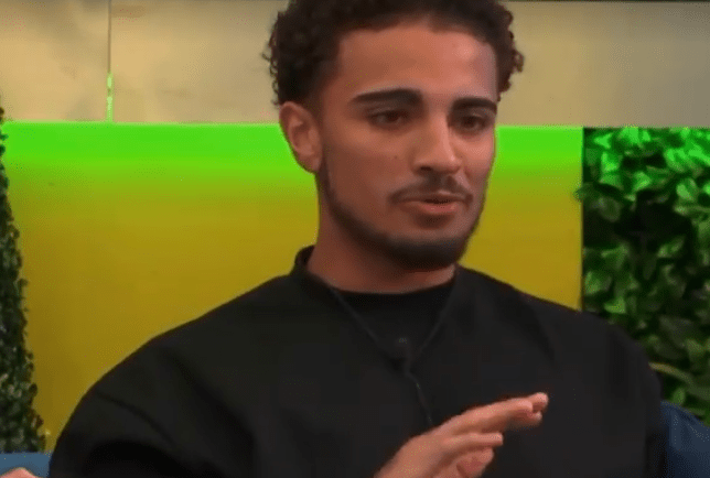 A young man with curly hair and a mustache gestures with his hand.