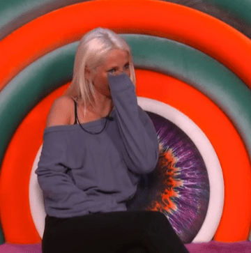 A blonde woman crying, with her hand covering part of her face, against a brightly colored background.