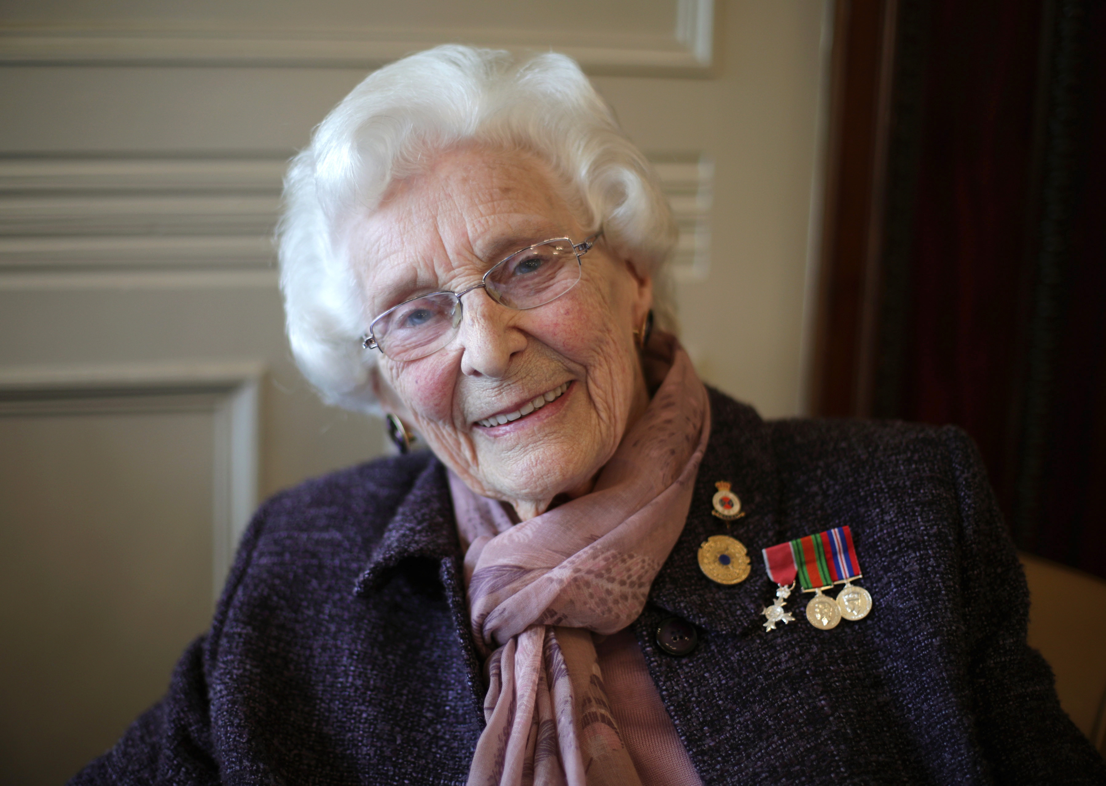 Bletchley Park veteran Betty Webb smiling at the camera, wearing a purple jacket with medals pinned to it and a pink scarf.