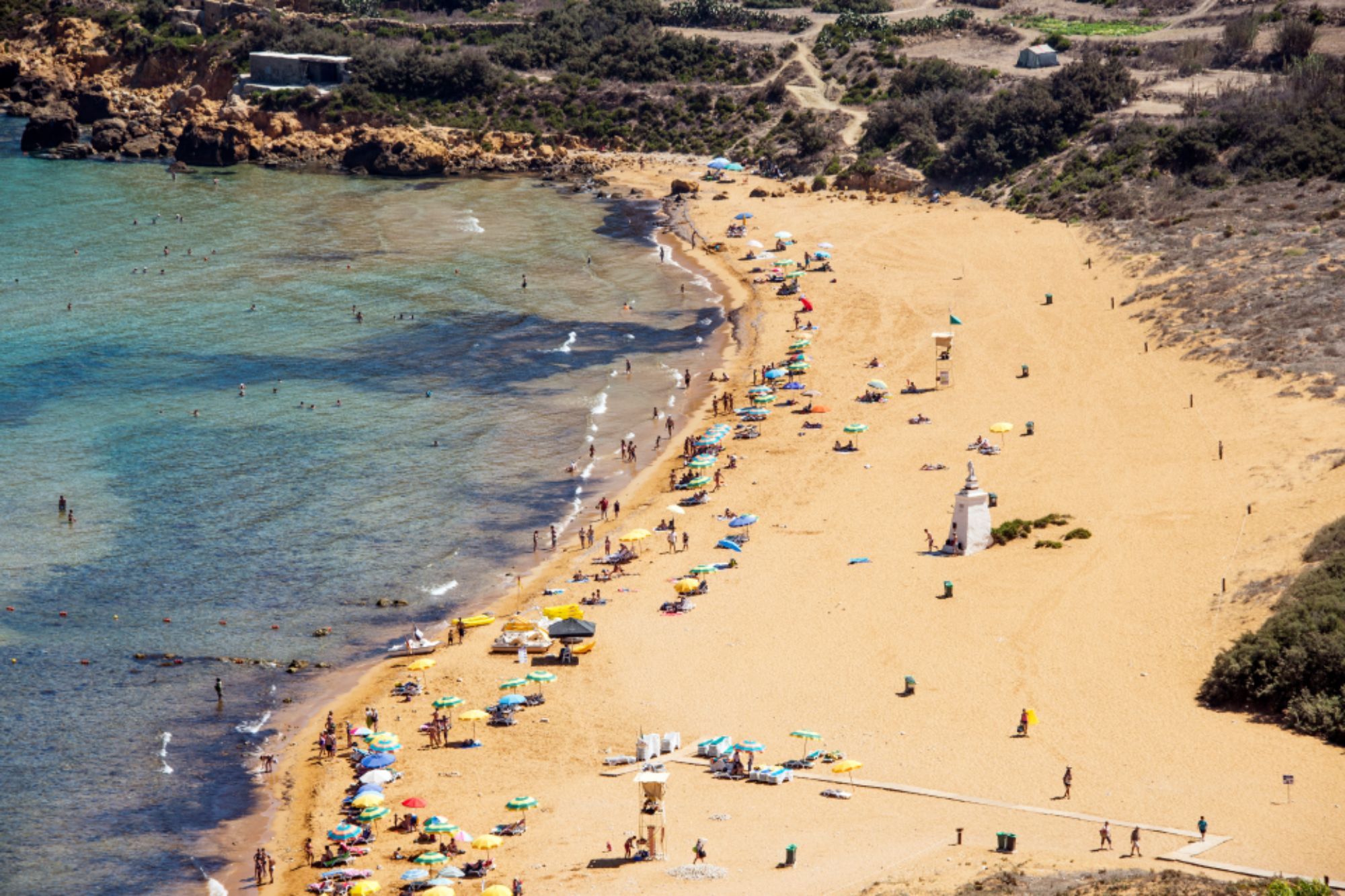An image collage containing 1 images, Image 1 shows Aerial view of Ramla Bay beach in Gozo, Malta, showing a sandy beach with many people, colorful umbrellas, and clear blue water