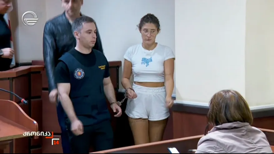 A woman in white shorts and t-shirt, handcuffed, stands in a courtroom next to a guard.