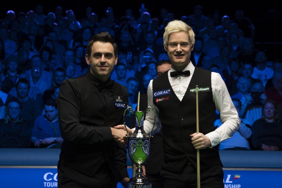 Ronnie O'Sullivan and Neil Robertson shaking hands with a trophy between them.