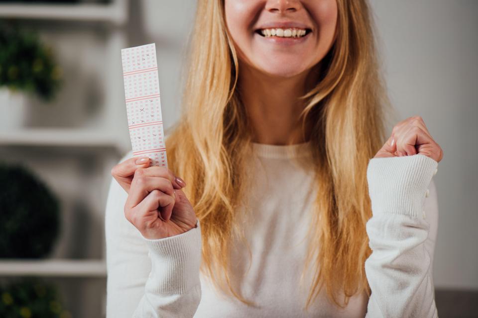 A smiling woman with blonde hair holds a lottery ticket with winning numbers and makes a winner gesture with her other hand.
