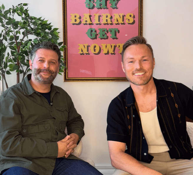 Two men seated, smiling, with a colorful poster behind them.