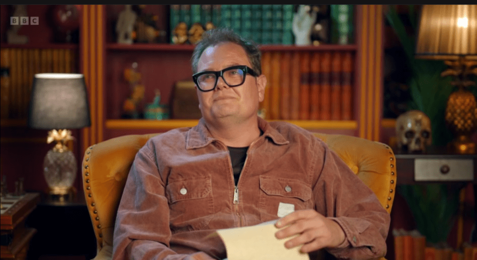 Alan Carr in glasses and a brown jacket sits in a yellow armchair, holding papers.