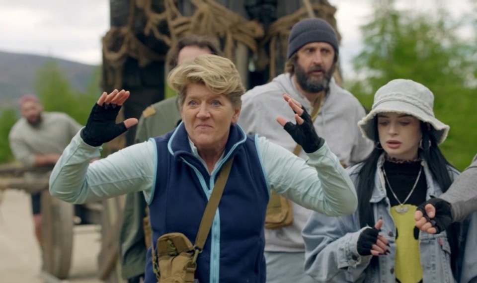 A woman with short blonde hair and a blue vest with black fingerless gloves throws her hands up in frustration.