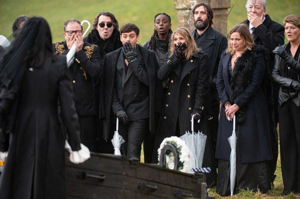 A group of people dressed in black, some holding umbrellas, stand over a coffin with expressions of shock and grief.